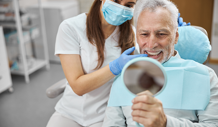 Older man in a dental chair holding up a hand mirror and looking at his brand new dental implants from Riverwind Dental in Richmond, VA