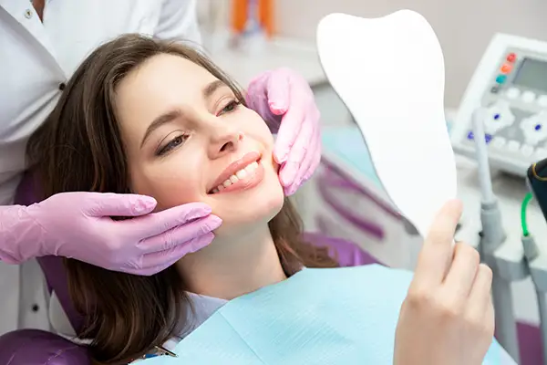 A smiling woman holding a tooth-shaped mirror while a dentist adjusts her face at Riverwind Dental in Henrico, VA