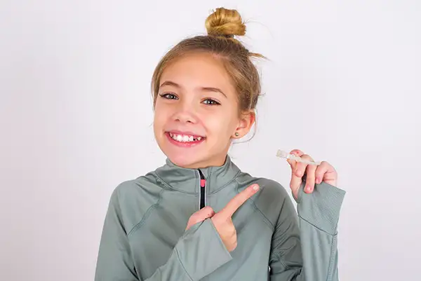 Young girl in a gray sweatshirt smiling and pointing to a clear aligner she is holding at Riverwind Dental in Richmond, VA