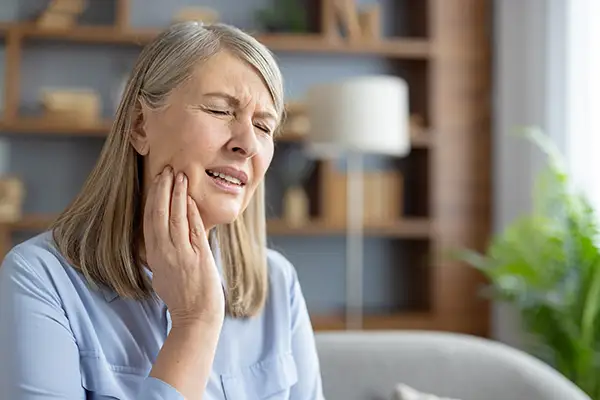 Elderly woman sitting at home, holding her jaw and grimacing in pain, indicating a dental emergency.