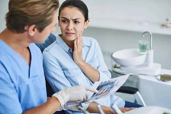 A dentist in scrubs discussing an X-ray with a female patient holding her jaw in pain at Riverwind Dental in Henrico, VA A dentist in scrubs discussing an X-ray with a female patient holding her jaw in pain at Riverwind Dental in Henrico, VA